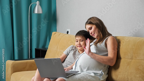 Pregnant woman and boy talking by video call on laptop while sitting on couch.