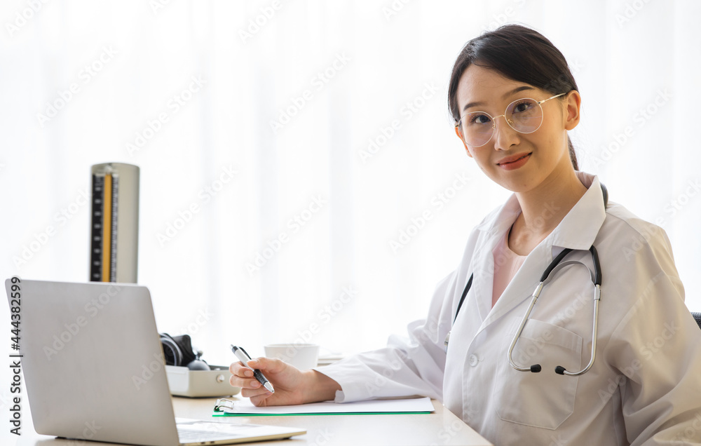 Young asian optometrist who wear cute eyeglasses in white uniform sit in the office clinic in hand holding pen and touch laptop. Seems busy but still have beautiful smile to camera man. Young asian optometrist who wear cute eyeglasses in white uniform sit in the office clinic in hand holding pen and touch laptop. Seems busy but still have beautiful smile to camera man.