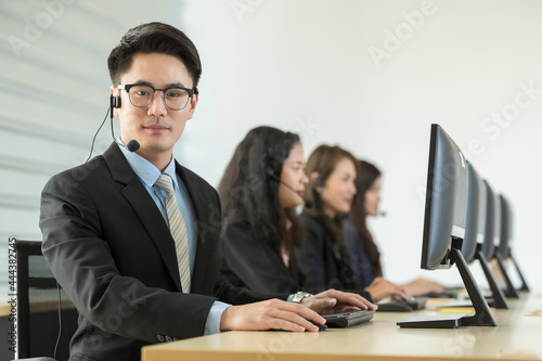 Cheerful Asian man in headset smile with cheerful and happy while working on computer at desk with female coworkers in help desk office. Sevice mind and ready for marketing support concept