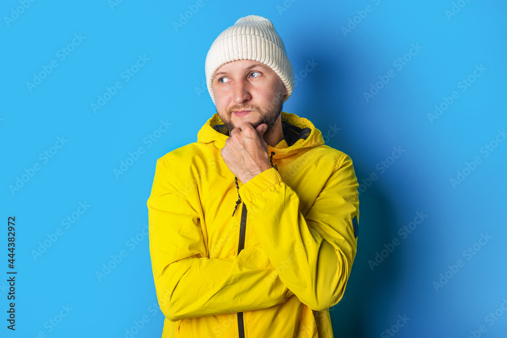 A pensive young man in a yellow jacket looks to the side while holding his hand under his chin on a blue background