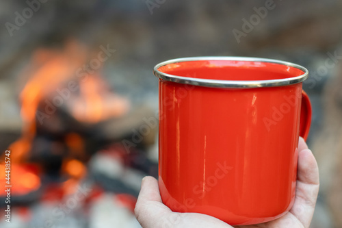Camping lifestyle concept. Hands holding red enamel mug near a campfire.