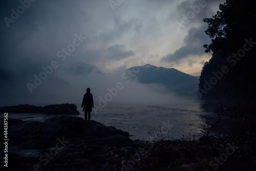 Alone human silhouette in white haze against the mountains and river. Thick fog in the evening twilight. Mysterious atmosphere. Reflection, meditation.