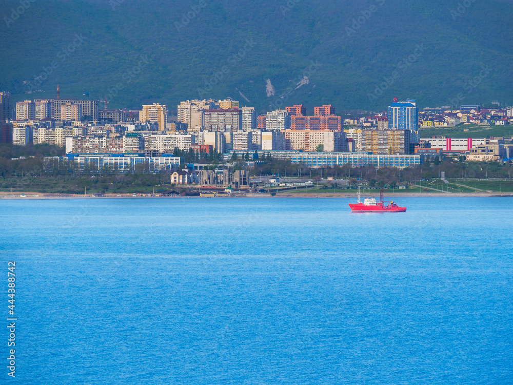 A red bright fire vessel with a crane goes through the black Sea in the Tsemesskaya Bay. Cargo ship on the background of the houses of the city of Novorossiysk.