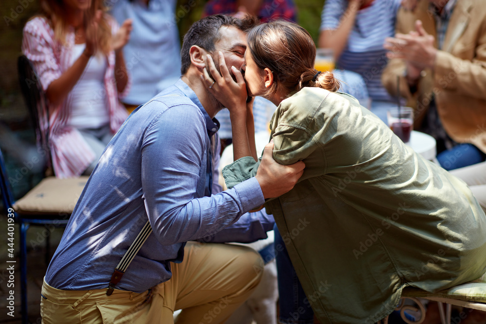 Fototapeta premium A young man kissing his girlfriend after asking to marry him at a party in a bar with friends. Leisure, bar, love, outdoor