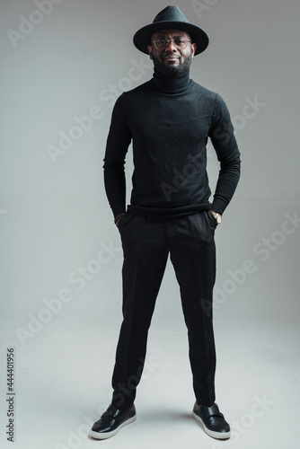 Full-length photo. Handsome african american man in glasses in a hat and black clothes stands with hands in pockets