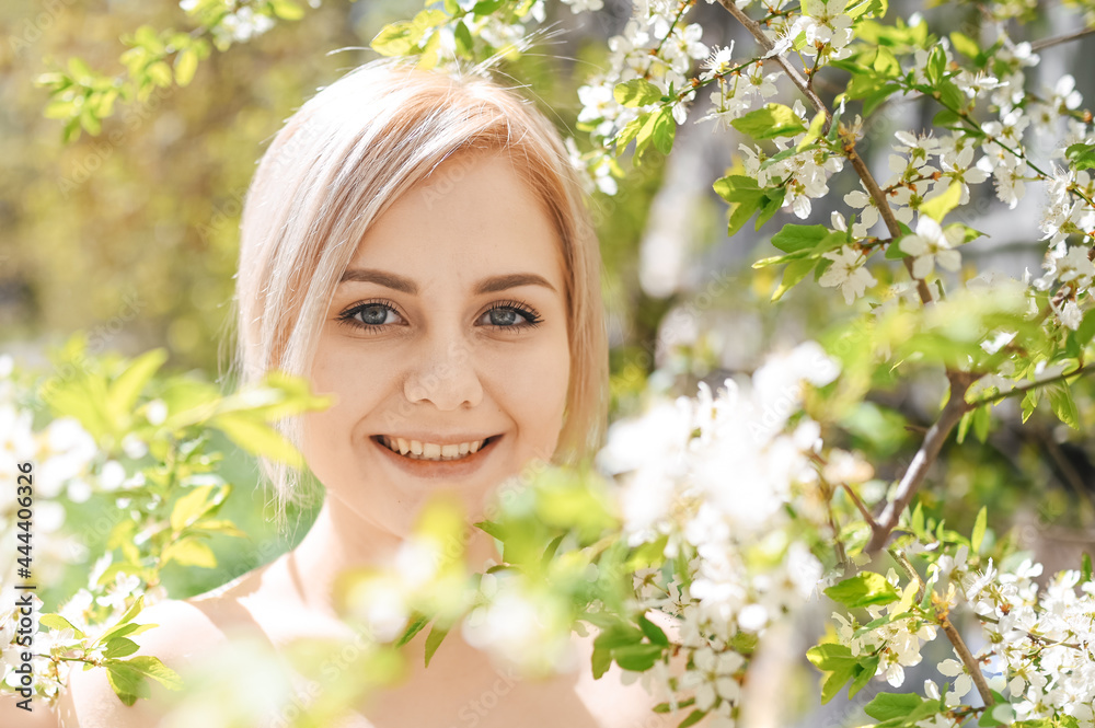 Fototapeta premium Young beautiful happy smiling blonde woman posing in blooming spring garden with white flowers. Blooming apple or cherry tree. Concept of natural cosmetics, youth, healthy skin and teeth