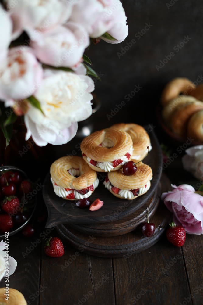 Custard rings with curd cream and berries on a dark table with a bouquet of peonies. Still life with flowers and sweets.