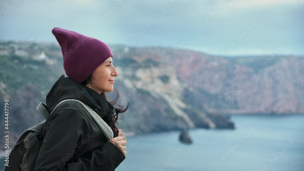 Pleasant hipster woman travel backpacker standing high cliff peak at windy weather enjoying nature