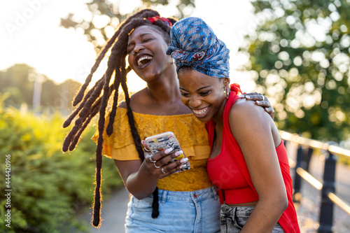 Two female friends with smartphone, smiling outdoors
