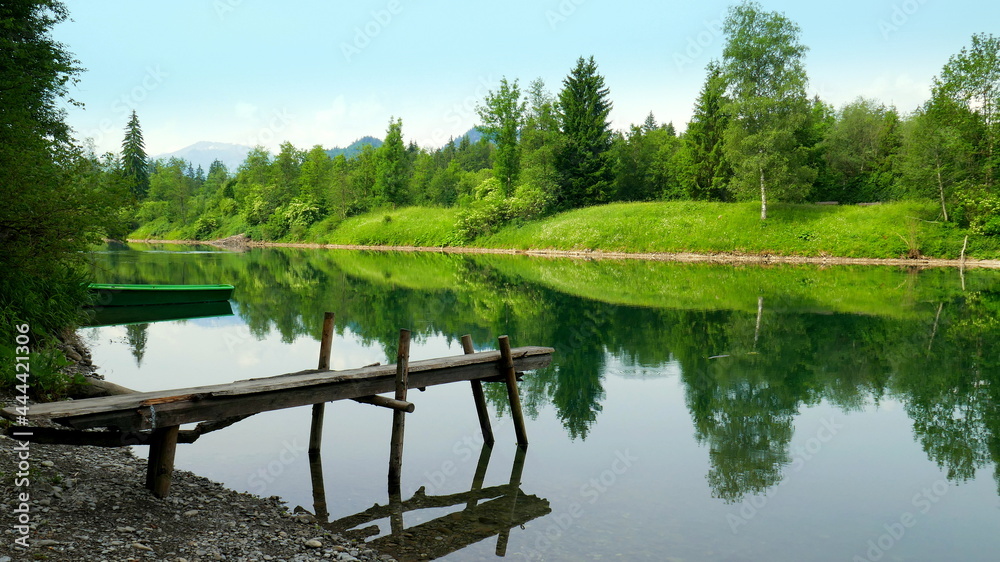 idyllischer Auwaldsee bei Fischen im Allgäu mit Boot und Bootssteg und Spiegelung von Wald und Wiesen 