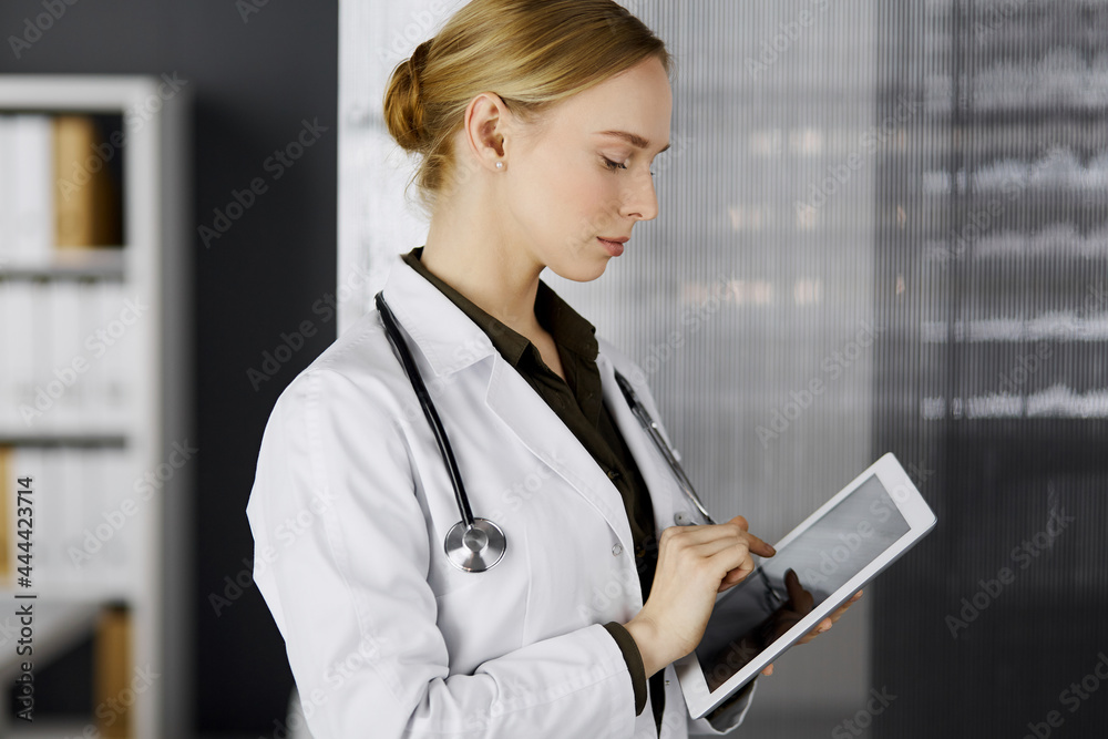 Friendly smiling female doctor using clipboard in clinic. Portrait of friendly physician woman at work place. Perfect medical service in hospital
