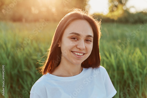 Portrait of young adult smiling woman wearing white clothing looking directly at camera with happy expression, posing in green meadow on sunset or sunrise.