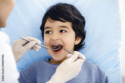 Cute arab boy sitting at dental chair with open mouth during oral checking up with doctor. Visiting dentist office. Stomatology concept