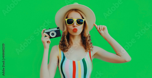 Summer portrait of attractive young woman with retro camera wearing a straw hat blowing her lips on green background