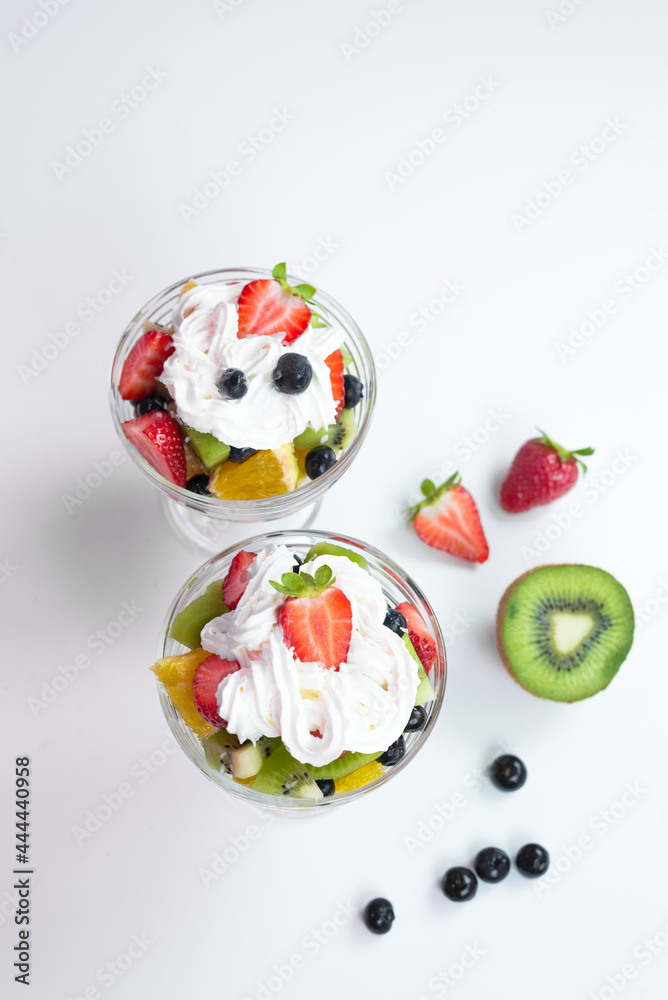 pieces of fresh fruits and berries with whipped cream and chocolate in a transparent bowl on a white table. 