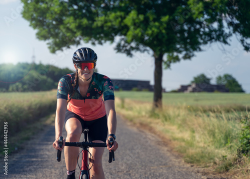 Fototapeta Naklejka Na Ścianę i Meble -  Frontal photo of a young woman cyclist, smiling, riding her road bike, in the middle of nature, illuminated by sunlight. She is wearing a helmet, sunglasses, and a pink cycling outfit.