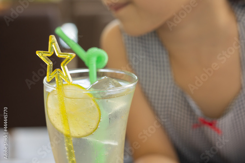 Close up and selective focus shot of lemonade soda in cocktail glass with fancy straw and lemon slice decoration in blurred background of adorable kid girl drinking for her refreshment.
