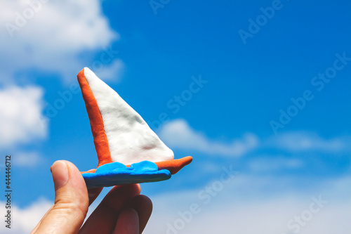 On the left, fingers are holding a brown boat sculpted from plasticine with a white sail and a blue wave against a blue sky with white clouds, close up 