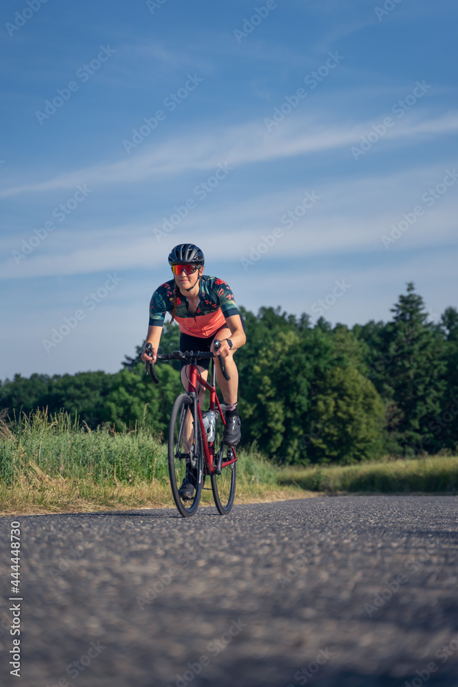 Vertical Angled photo, of a young woman professional cyclist, riding speed her road bike, on a paved road amidst nature, illuminated by sunlight. Sport Equality concept.