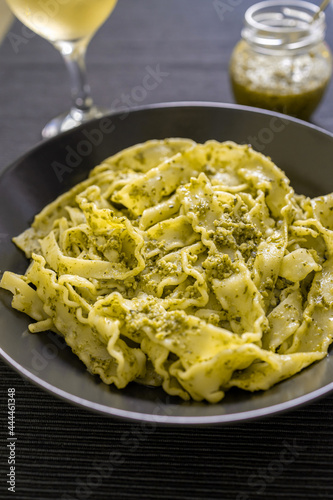 Pasta Mafaldine with tofu pesto sauce served on a dark plate. Jar with pesto sauce and a glass of wine on textile placemat.