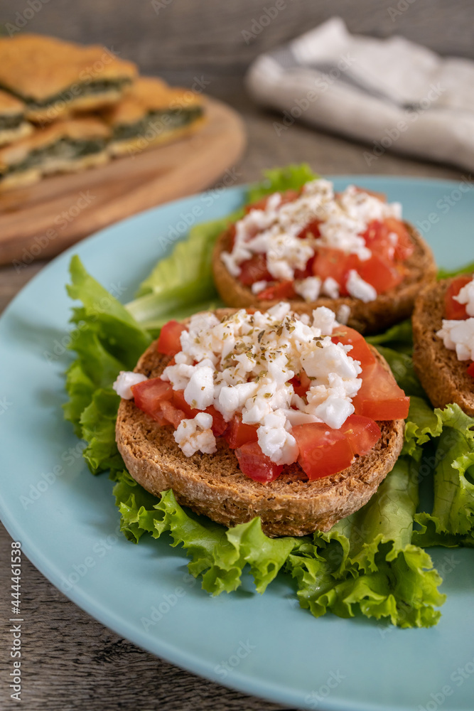 Burley rusks with chopped tomatoes and feta cheese served on lettuce leaves. Greek “Dakos” traditional food recipe with greens pie on wooden cutting board - half plate side view.