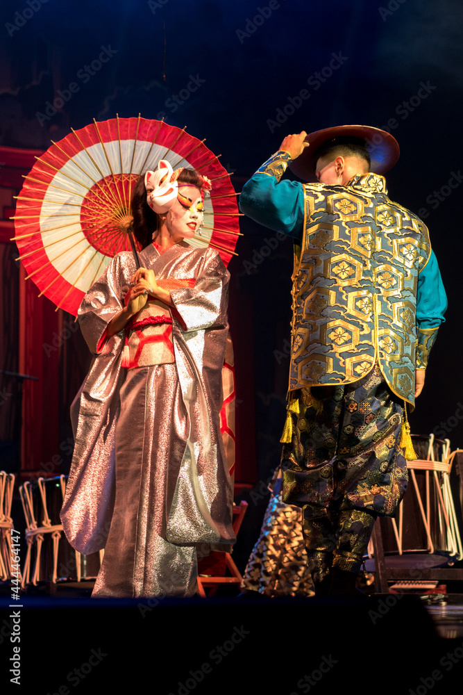 Japanese woman geisha in traditional kimono and  meets and Kino Kitsune fox mask greets man in a dark background. Traditional Japanese performance.