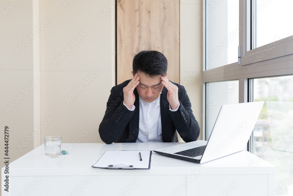 young asian office worker suffering from headache at work in the office drinks medicine pill with water. Sick man in a suit at the computer indoors with severe pain ache uses the drug in workplace young asian office worker suffering from headache at work in the office drinks medicine pill with water. Sick man in a suit at the computer indoors with severe pain ache uses the drug in workplace