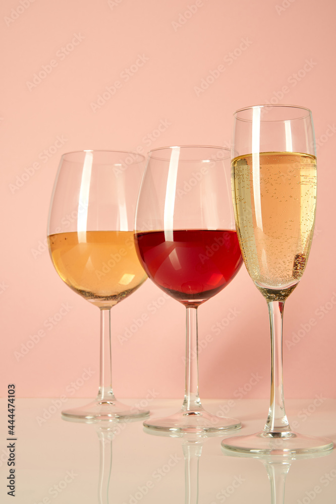 Three glasses of different shapes and containers for wine stand on a white glossy table against a pink background. Glasses with white and red wine
