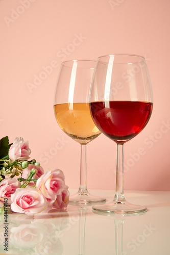 Two wine glasses are on a glossy table next to roses. Glasses with white and red wine on a pink background