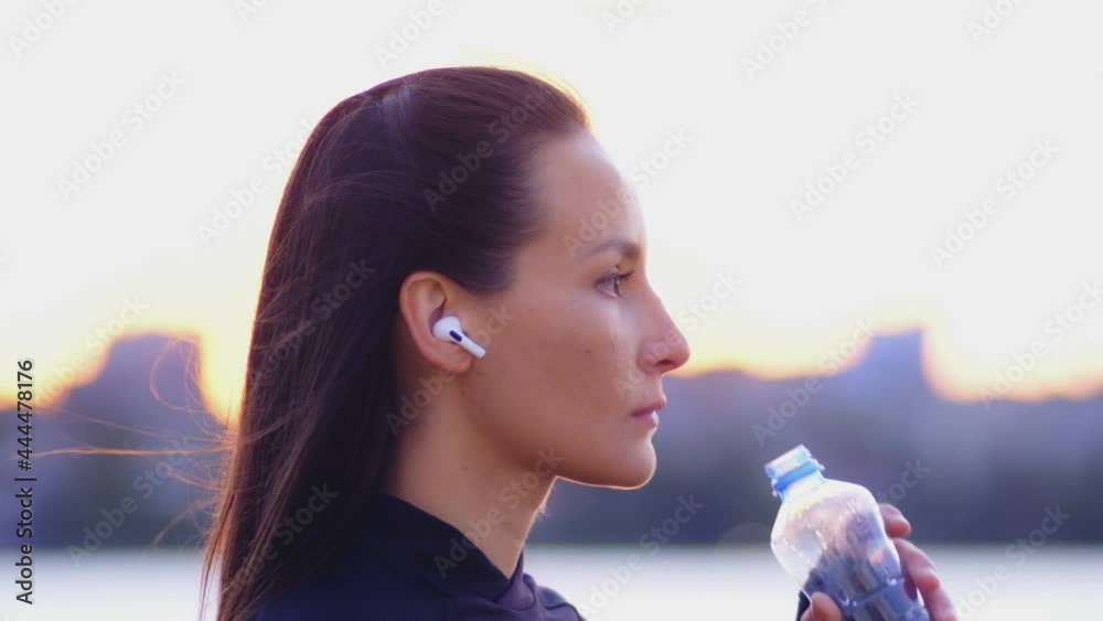 Asian young beautiful brunette woman in sportswear and wireless earphones drinks water from a plastic bottle. Her hair is blowing in the wind. The girl confidently looks straight ahead Asian young beautiful brunette woman in sportswear and wireless earphones drinks water from a plastic bottle. Her hair is blowing in the wind. The girl confidently looks straight ahead