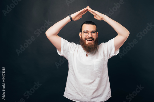 Portrait of satisfied happy bearded man in casual making house symbol, roof gesture over head and smiling, dark background