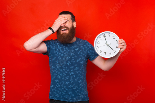 Portrait of bearded younfg man holding clock and keeping facepalm gesture, expressing regret of missed deadline photo