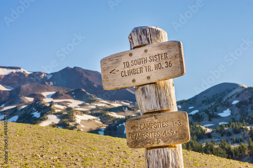 Wooden signpost guides hikers along a trail to a distant volcanic mountain summit peak.