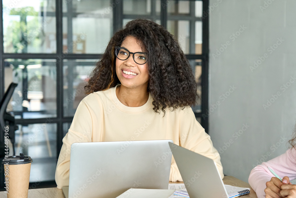 Young happy smiling African American girl student in eyeglasses at desk working using pc laptop computer at modern office university college campus listening lesson class. Education concept. Young happy smiling African American girl student in eyeglasses at desk working using pc laptop computer at modern office university college campus listening lesson class. Education concept.