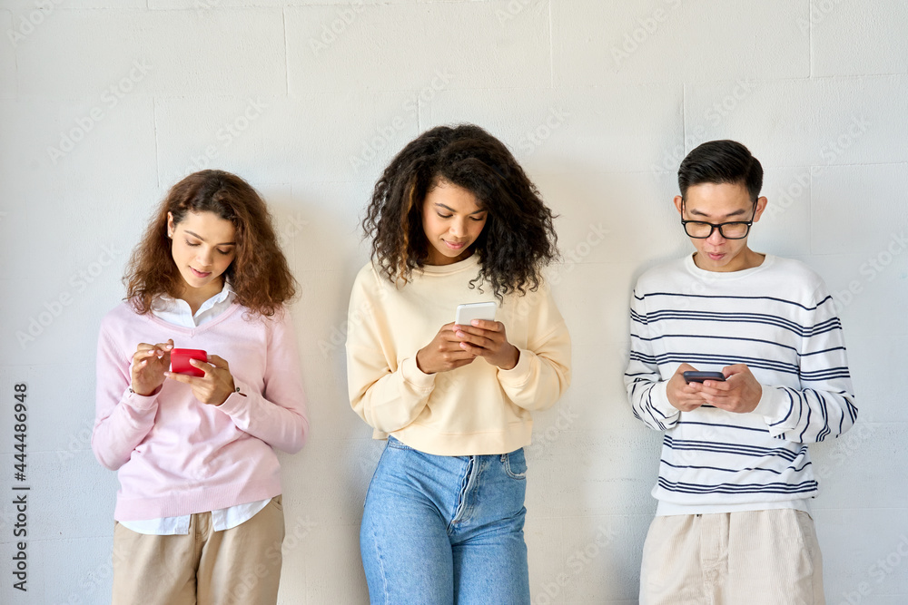 Young teenage cool multiethnic group of gen z high school college students standing on white wall background holding mobile cell phones looking at smartphones using apps. Social media concept Young teenage cool multiethnic group of gen z high school college students standing on white wall background holding mobile cell phones looking at smartphones using apps. Social media concept