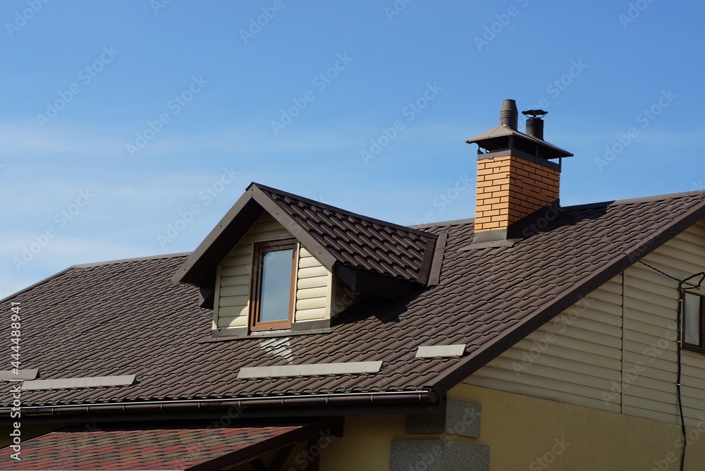 part of a private house from an attic with a window under a brown tiled roof with a brick chimney on the background of a blue house