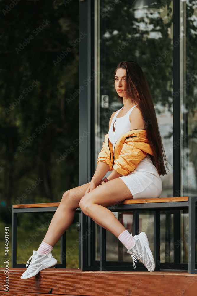 Outdoor close up portrait of young beautiful woman in white dress and yellow coat sitting on bench in  park.  Autumn walking concept.