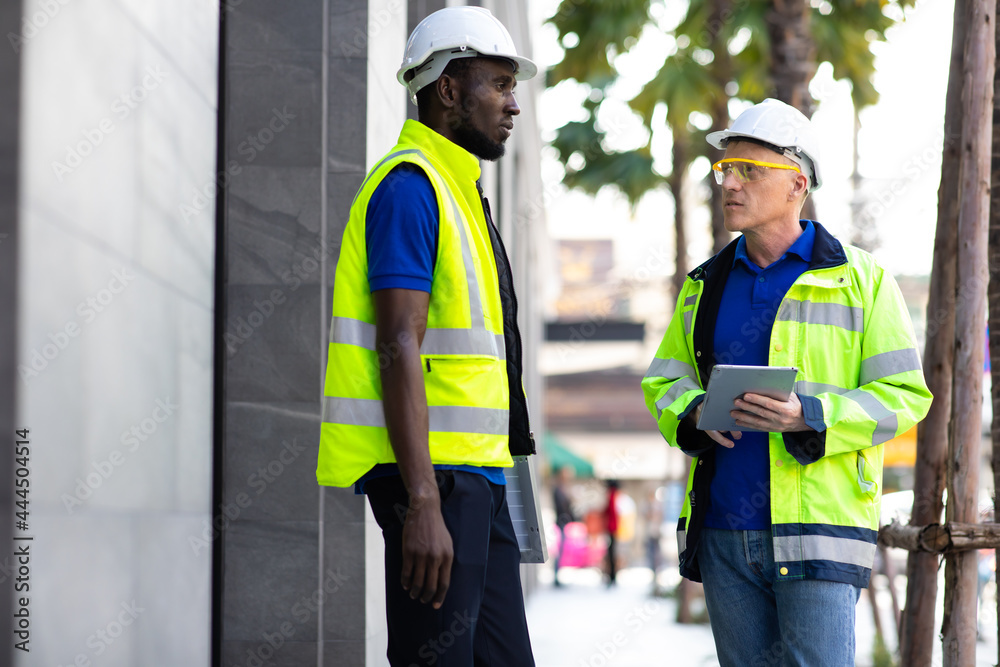 Two engineers African american engineer and caucasian electrician wearing white hard hat walk in new building holding solar panel on hand and Discuss Work Two engineers African american engineer and caucasian electrician wearing white hard hat walk in new building holding solar panel on hand and Discuss Work