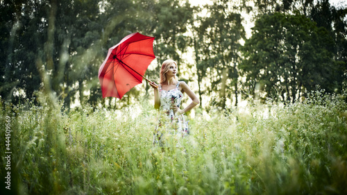A young slender woman walks on nature in a light summer sundress with a floral print under a red umbrella