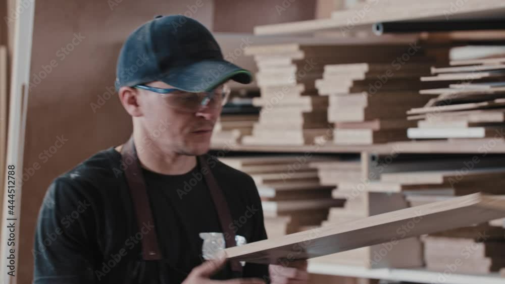 Man in a carpentry workshop takes a wooden piece from the shelf and blowing dust from it - looking in the camera Man in a carpentry workshop takes a wooden piece from the shelf and blowing dust from it - looking in the camera