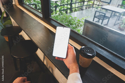 Close up image of a person using a mock up smartphone with blank screen in a coffee shop