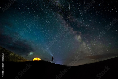 star fall on the background of a night sky full of stars with a milky way and a silhouette of a man and an orange tent on the horizon