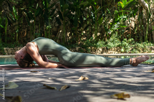 Young woman with blonde hair in Matsyasana yoga pose next to blue swimming pool and garden photo