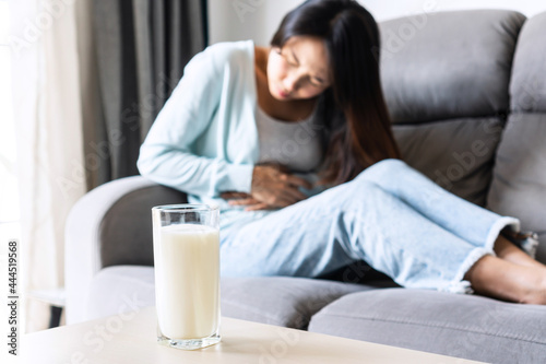 Unhappy young Asian woman having bad stomach ache with glass of milk on table. Lactose intolerance, food allergy concept.