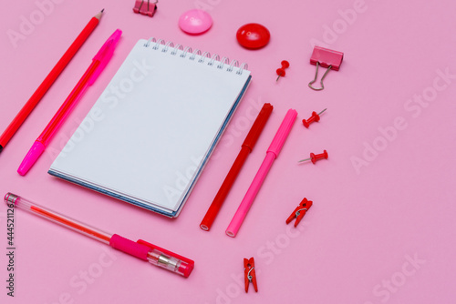 Markers of different pink shades with white notepad on pink background, top view. Pens and pencils are laid out around the notebook close-up