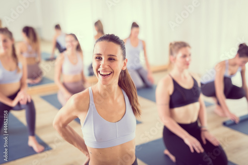 Portrait of a cheerful beautiful young yoga instructor relaxing after giving yoga class to large group of sporty attractive people. Healthy active lifestyle, working out indoors in gym.