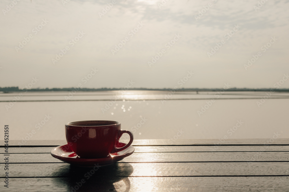 Red cup of hot latte coffee on wooden table at sunrise.