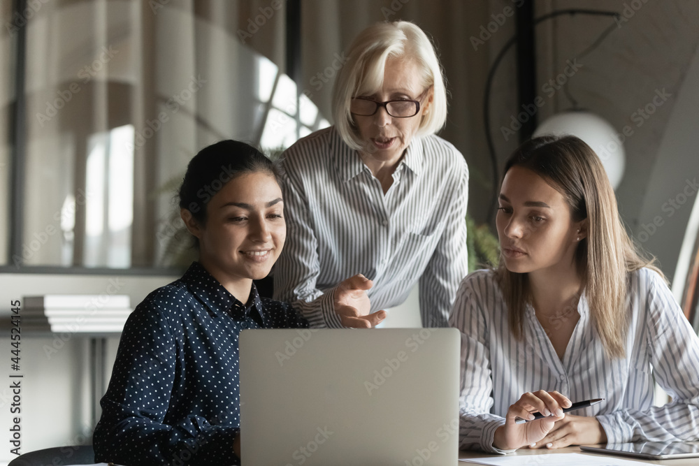 Corporate teacher showing and explaining work data to student girls, pointing at laptop screen, speaking. Mentor training interns. Elder coach giving advice to diverse office apprentices Corporate teacher showing and explaining work data to student girls, pointing at laptop screen, speaking. Mentor training interns. Elder coach giving advice to diverse office apprentices