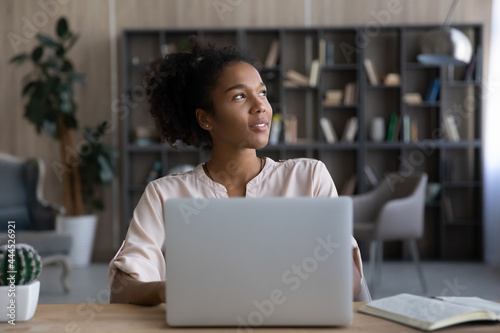 Dreamy African American woman looking to aside, distracted from laptop, sitting at work desk, happy young female dreaming about good future or new opportunities, pondering strategy, planning project photo