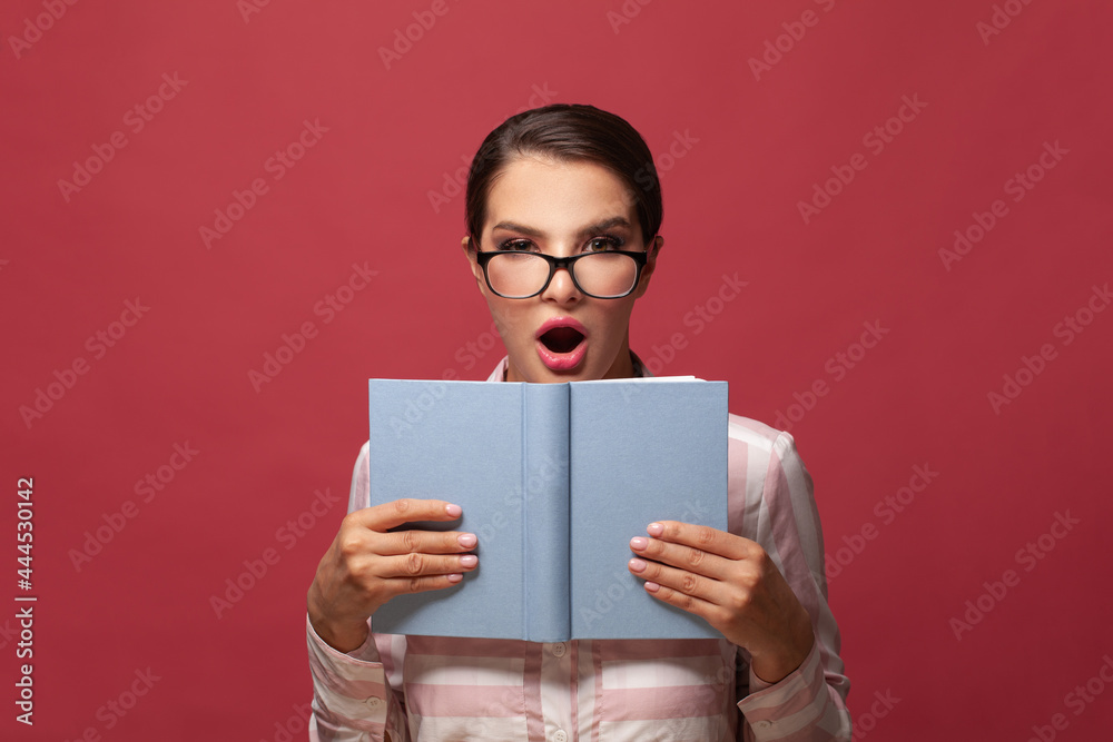 Portrait of smart surprised woman in glasses holding book with empty cover posing isolated on red wall background. Mock up copy space. People sincere emotions lifestyle concept Portrait of smart surprised woman in glasses holding book with empty cover posing isolated on red wall background. Mock up copy space. People sincere emotions lifestyle concept